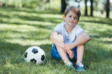 cute little boy sitting on grass with soccer ball and looking at camera in park