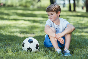 cute little boy sitting on grass and looking at soccer ball