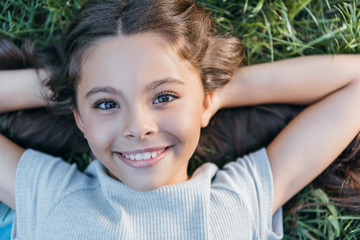 top view of happy child lying with hands behind head on grass and smiling at camera