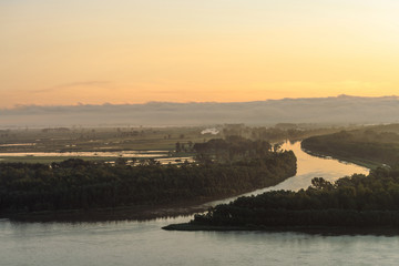 Broad river flows along shore with forest with mist. Channel of river flows around island. Orange glow at dawn reflected on water. Colorful morning mystical atmospheric landscape of majestic nature.