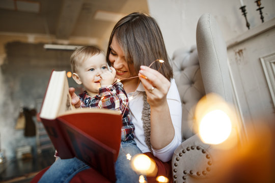 Mother is sitting and holding babu on lap. She is looking at child and smiling. Young woman is holding garland and book. Baby is looking at garland with interest. Child is smiling.