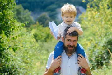Father, son and plane in nature. Bearded parent holds on shoulders cheerful cute boy with paper airplane in hands. Walking and happy childhood, hobby aviation and family travel to airport