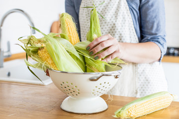 Woman holding colander with yellow corn cobs. View on white modern scandinavian style kitchen
