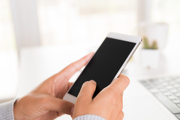 Woman hand holding white mobile phone on a table with a laptop in office.