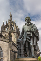 Fototapeta na wymiar Edinburgh, Scotland, UK - June 14, 2012: Adam Smith bronze statue on market square in front of brown stone Saint Gilles Cathedral under blue sky with clouds. 
