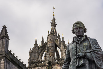 Edinburgh, Scotland, UK - June 14, 2012: Adam Smith bronze statue on market square in front of brown stone Saint Gilles Cathedral crown tower under gray silver sky. 