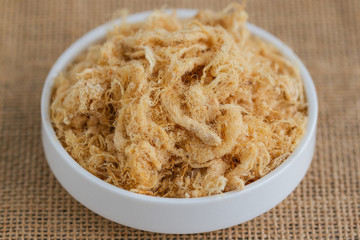 Homemade sweet and soft dried shredded pork or pork floss on white plate put on sack in close up view macro concept to present texture. Thai style delicious food for cooking or bakery.