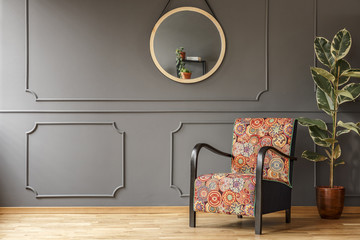 Ficus next to patterned armchair against grey wall with molding and mirror in flat interior. Real photo
