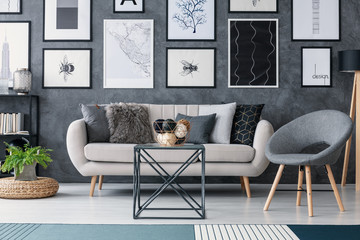 Grey chair next to sofa and table in modern living room interior with plant and posters. Real photo