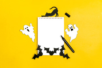 Crafts for the concept celebration of Halloween - figures of a black bat,cat,ghost cut from paper from stencil around a white notepad, felt-tip pen on yellow background Place for text Mock up Top view