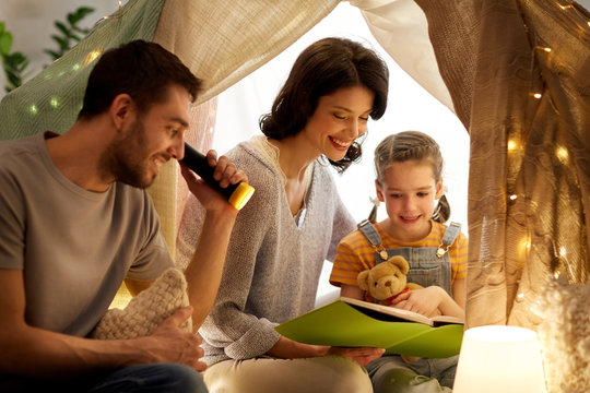 family, hygge and people concept - happy mother, father and little daughter reading book with torch light in kids tent at night at home