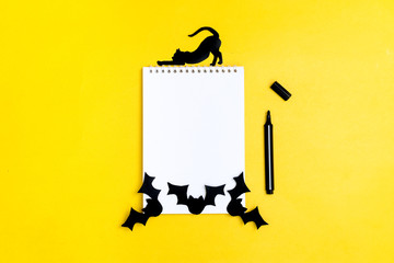 Crafts for the concept celebration of Halloween - figures of a black bat,cat,ghost cut from paper from stencil around a white notepad, felt-tip pen on yellow background Place for text Mock up Top view
