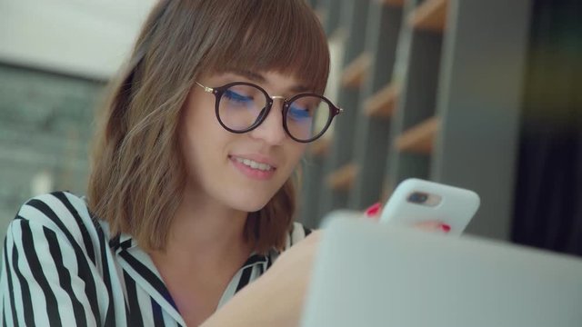 Stylish young girl in a striped shirt and glasses is sitting in a bright room with a phone in hands and smiling. A young successful woman looks into the phone and smiles. Lightness, good mood, youth