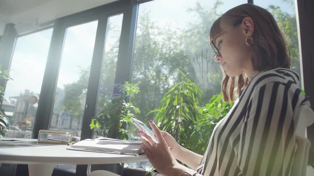 A slender brunette woman in a black and white striped blouse sits alone in a cafe with large panoramic windows overlooking the city and uses a smartphone. Modern woman and gadget.