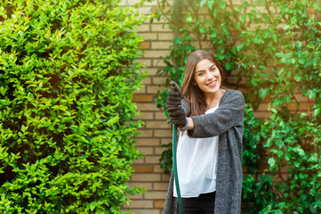 Naklejka premium Smiling young woman with garden hose watering her home backyard with flowers, plants and vegetation. Gardening as hobby and leisure concept.