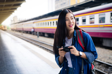 Obraz premium Young Asian woman smile holding camera in hand. Young passenger girl looking, waiting railway at train station. Asian backpack traveler girl smiling enjoy holding camera standing at train station.