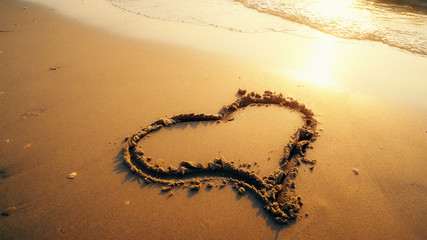 Hart shape drawn on beach sand is being washed by sea waves with sunset reflection