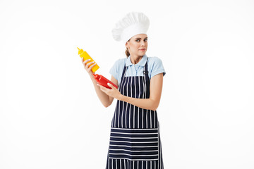 Young astonished woman chef in striped apron and white hat thoughtfully looking in camera with bottles of ketchup and mustard over white background isolated