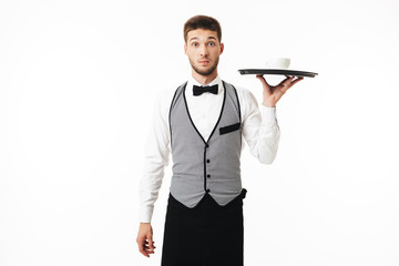 Young shocked waiter in uniform holding tray with cup of coffee while amazedly looking in camera over white background