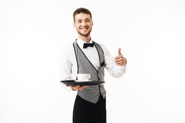 Young smiling waiter in uniform holding tray with cup of coffee in hand joyfully looking in camera...