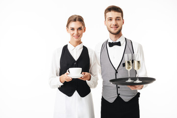 Young smiling waiter holding tray with glasses of champagne while pretty waitress holding cup of...