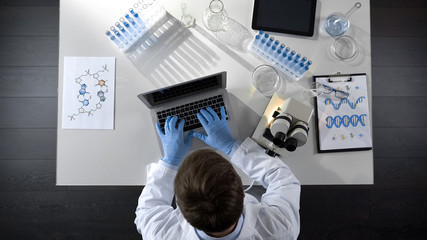 Lab worker viewing samples under microscope and typing results on laptop topview