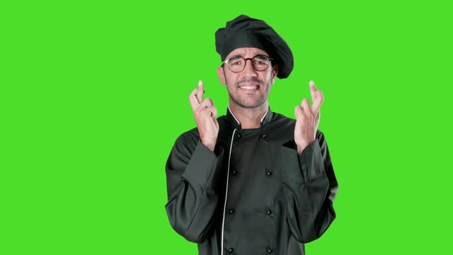 Worried young chef with a crossed fingers gesture against green background