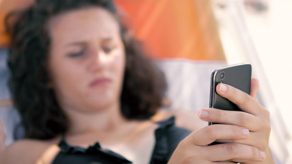 Obraz na płótnie Canvas Female teen fingers type message on smartphone lying on beach sunbed, cinematic dof