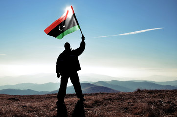 Successful silhouette man winner waving Libya flag on top of the mountain peak - obrazy, fototapety, plakaty