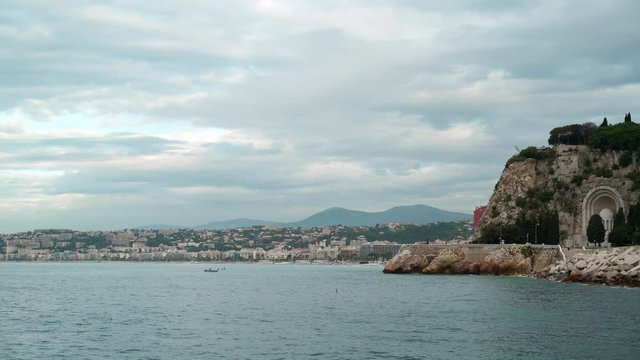 Coast of French resort of Nice on a windy and cloudy spring day. People are rowing in boats and fishing. Sea and sky. Monuments. Concept of travel. Right to left pan real time establishing shot