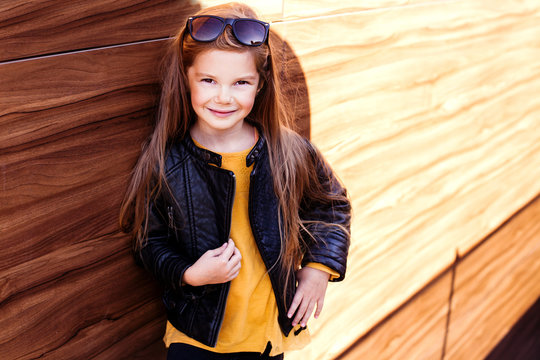 Smiling cool and looking sharp girl in sunglasses wearing black leather jacket standing on the wooden background. Street fashion. City style. Kids fashion. Autumn. 