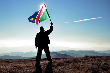 Successful silhouette man winner waving Namibia flag on top of the mountain peak