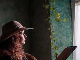 Portrait of a young woman with curly hair wearing a straw hat with natural light coming in through a window in a rural house