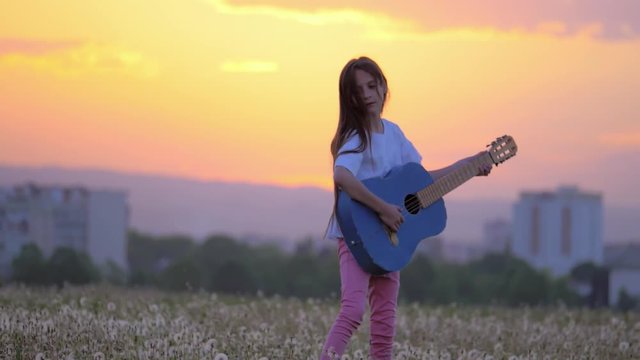 wonderful sunset little kid girl hipster playing the guitar like a pop star in a dandelions field . outdoors away from town buildings