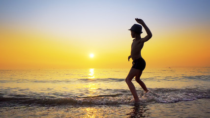 Fototapeta na wymiar Silhouette of boy with hat throwing stones skipping on sea water surface. Summer vacation concept with vibrant orange sky