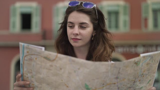 Attractive young woman tourist in purple sunglasses and straw hat looking around while holding large map standing in French city of Nice on sunny spring day. Right to left pan slow motion medium shot