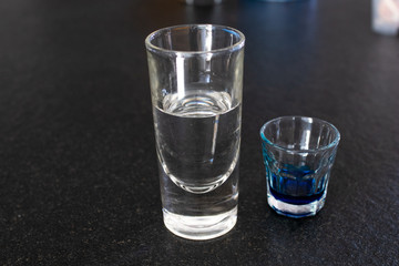 Tall transparent glass with water-like liquid with a small glass near it with blue liquid. Two glasses with different liquids on a black granite table