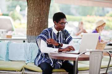 African man in traditional clothes and glasses sitting behind laptop at outdoor caffe.