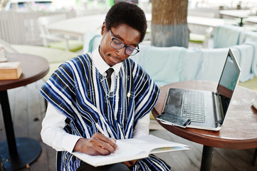 African man in traditional clothes and glasses sitting behind laptop at outdoor caffe and writing something on his notebook.