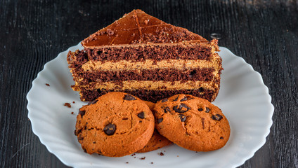 a piece of chocolate cake and cookies with chocolate on a white plate on a dark wooden background. Homemade baking.Close-up view.