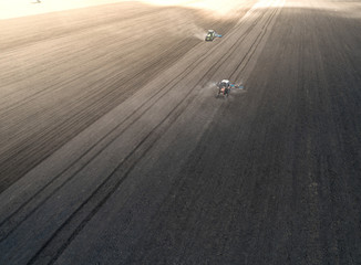 Aerial view of two blue tractors plows the earth in field on a summer day against a black earth...