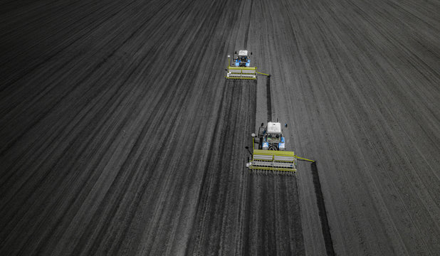 Aerial view of two blue tractors plows the earth in field on a summer day against a black earth background. Agriculture. Two tractors travel one after another along the black field