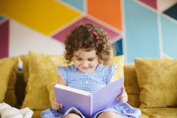 Fototapeta na wymiar Beautiful little smiling girl with dark curly hair in blue dress reading book on sofa at home