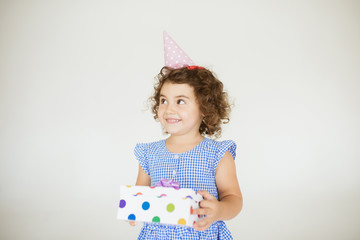 Pretty smiling baby girl with dark curly hair in blue dress and birthday cap happily looking aside while holding gift box over white background