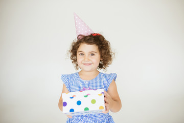 Beautiful smiling little girl with dark curly hair in dress and birthday cap happily looking in camera with gift box in hands over white background