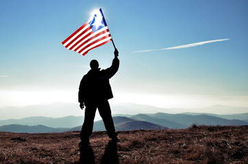 Successful silhouette man winner waving Liberia flag on top of the mountain peak - obrazy, fototapety, plakaty
