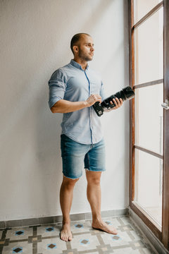 Brutal photographer in the blue shirt and blue jeans shorts stands on the floor of the flat with white walls holding his camera near the window in Spain 