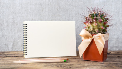 White empty notepad, notebook, wooden colored pencils and green cactus in brown pot with bow on wooden table on gray fabric background. Hipster eco natural mockup with copy space.