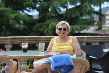Aged lady is sitting on terrace with glass in hands.