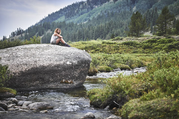 a young woman with curly hair relaxing on a giant boulder next to a mountain stream